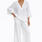 Woman wearing white linen co-ord set with V-neck top and wide-leg drawstring pants, styled against white flower wall.