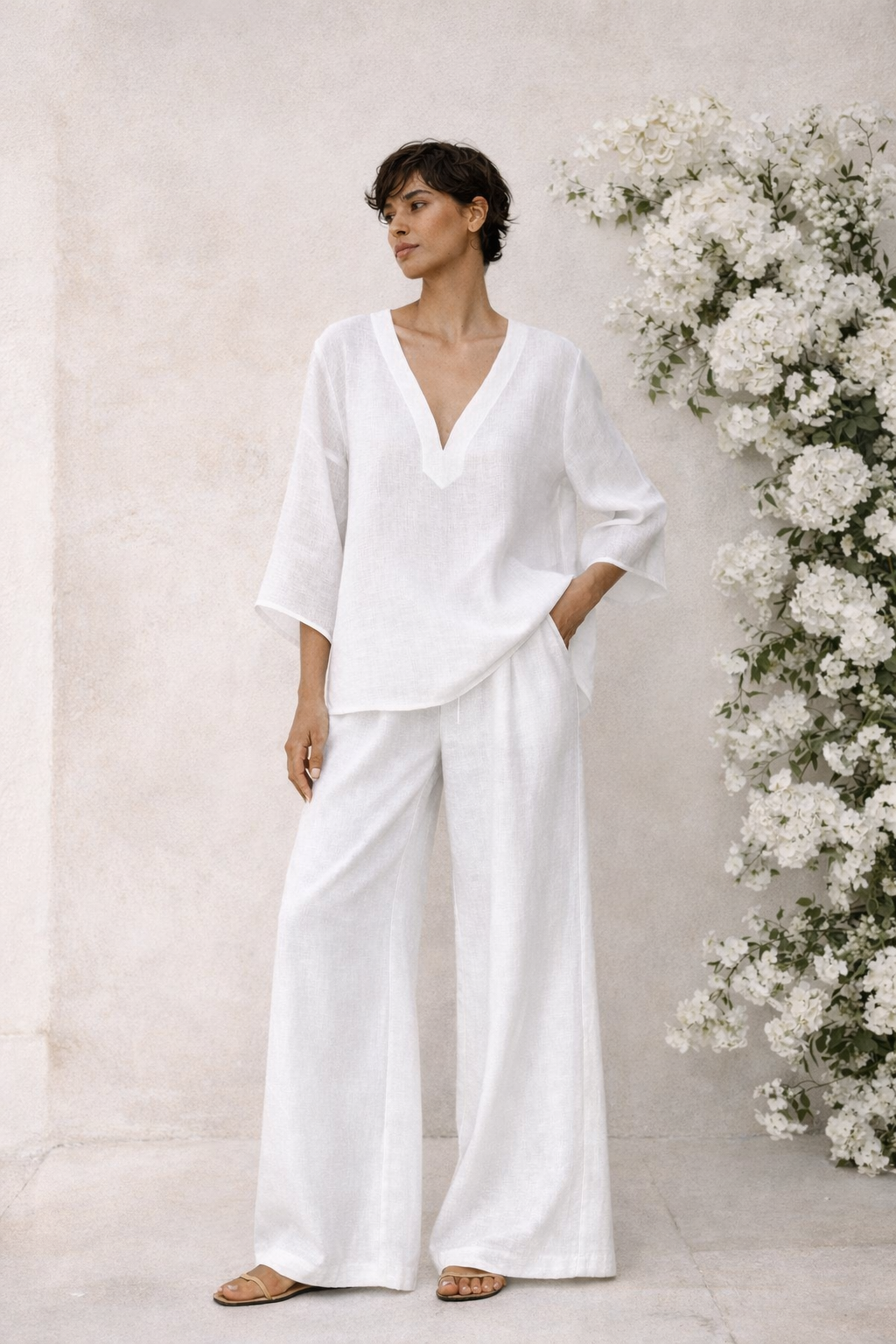 Lifestyle image of woman wearing white linen co-ord set styled against minimal wall and white floral background.